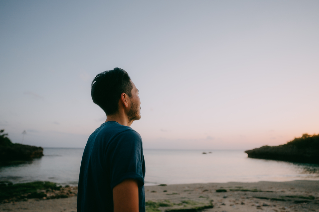 A person stands on a sandy beach, gazing at the calm ocean during sunset, evoking a sense of tranquility and reflection
