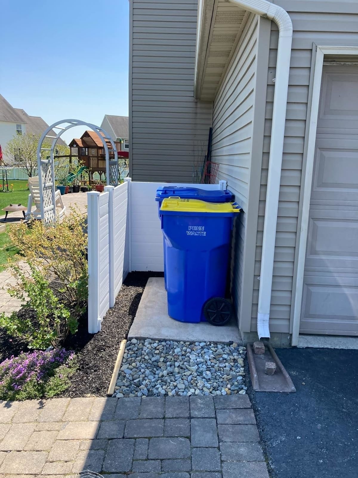 same reviewer's blue recycling bin with a yellow lid placed on a side path next to a garage, hidden by a privacy fence