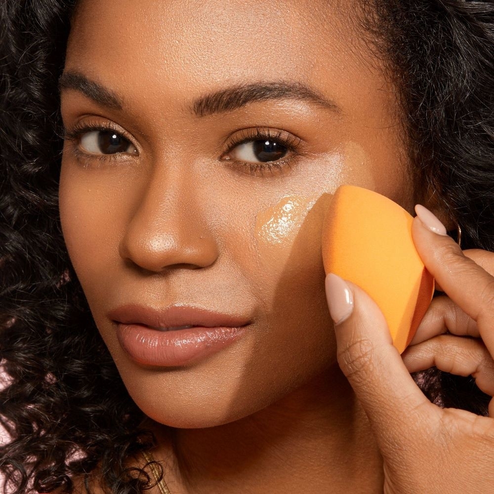 Person applying foundation to face with an orange makeup sponge. The focus is on makeup application technique