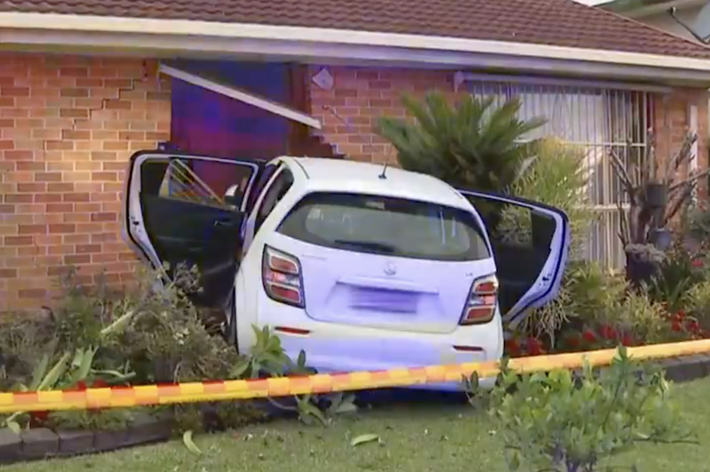 自動車が庭に突っ込み、家の壁に突き刺さっている事故現場。警察のテープが周囲を囲んでいる。