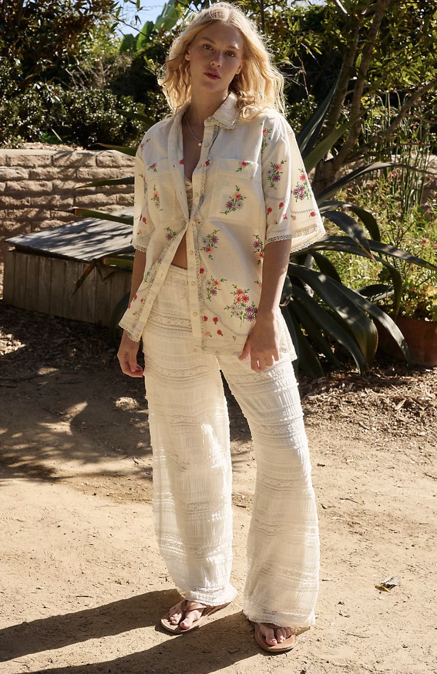 Orang yang mengenakan kemeja kancing bunga dan celana putih bertekstur berdiri di luar ruangan dalam pengaturan taman