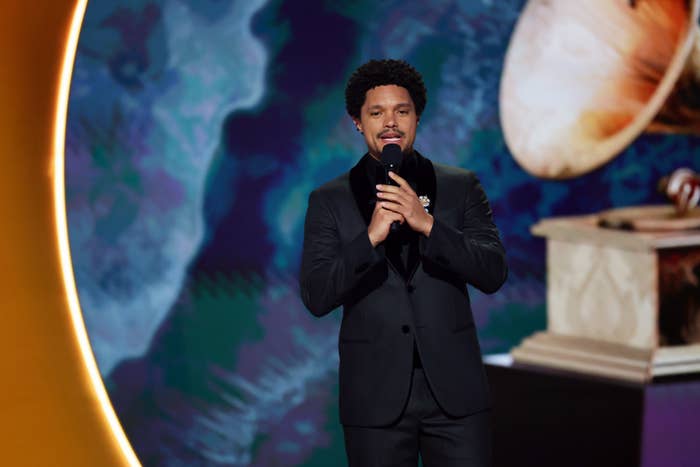 Person in a dark suit holding a microphone, standing in front of a large award statue backdrop