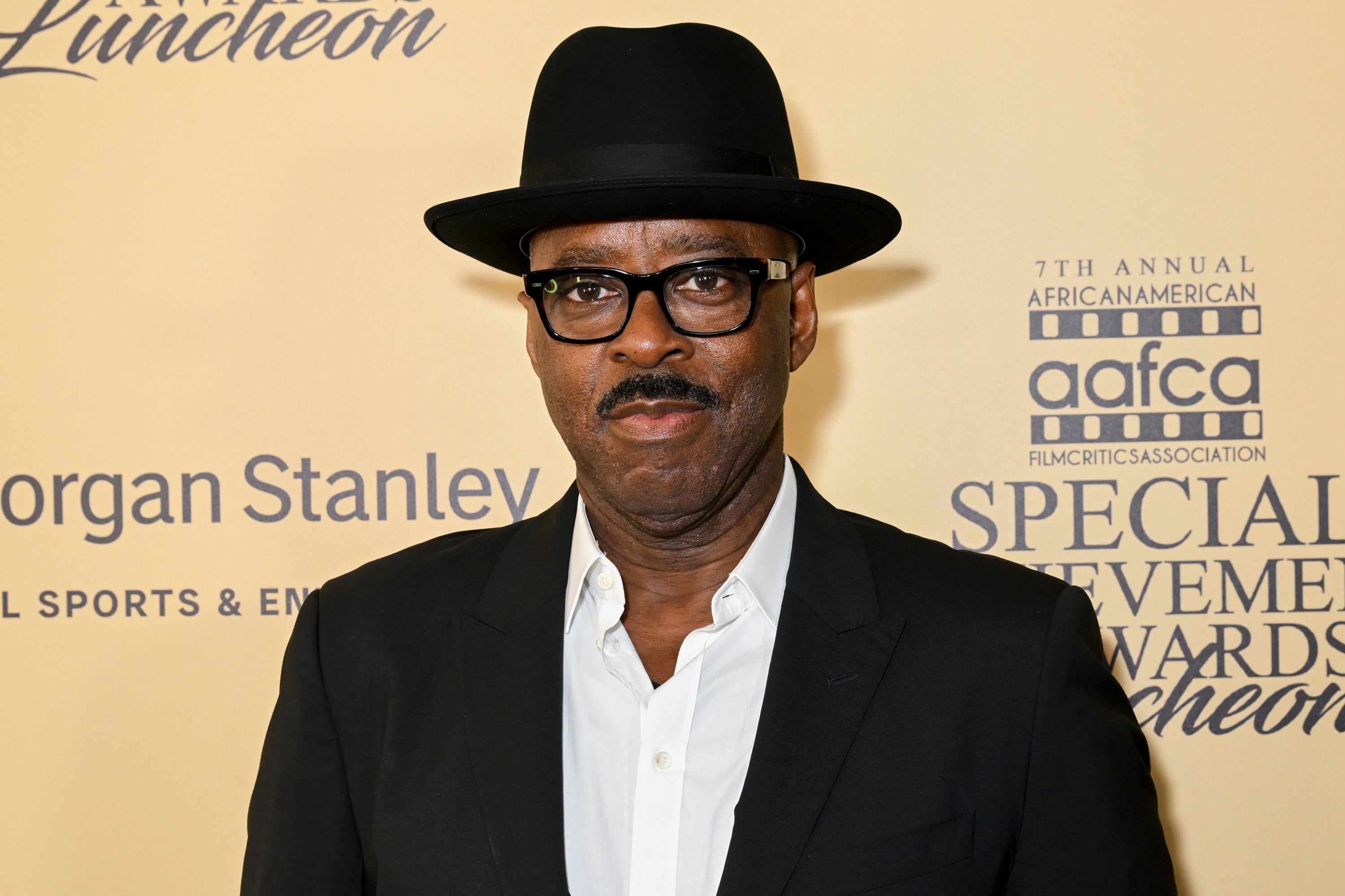 Person in a black suit and hat with a mustache stands in front of a backdrop at an awards event