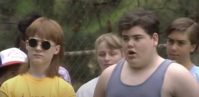 Group of teenagers stand outdoors, looking surprised. One wears sunglasses with long hair, another a tank top. Trees are visible in the background