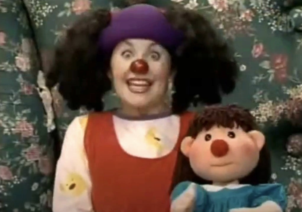 A person dressed as a clown with a red nose and purple hat smiles widely, holding a doll with a similar red nose and playful expression