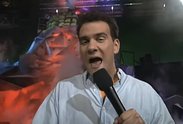 Person holding a microphone, speaking energetically in a studio with dramatic lighting and a smoke-filled background