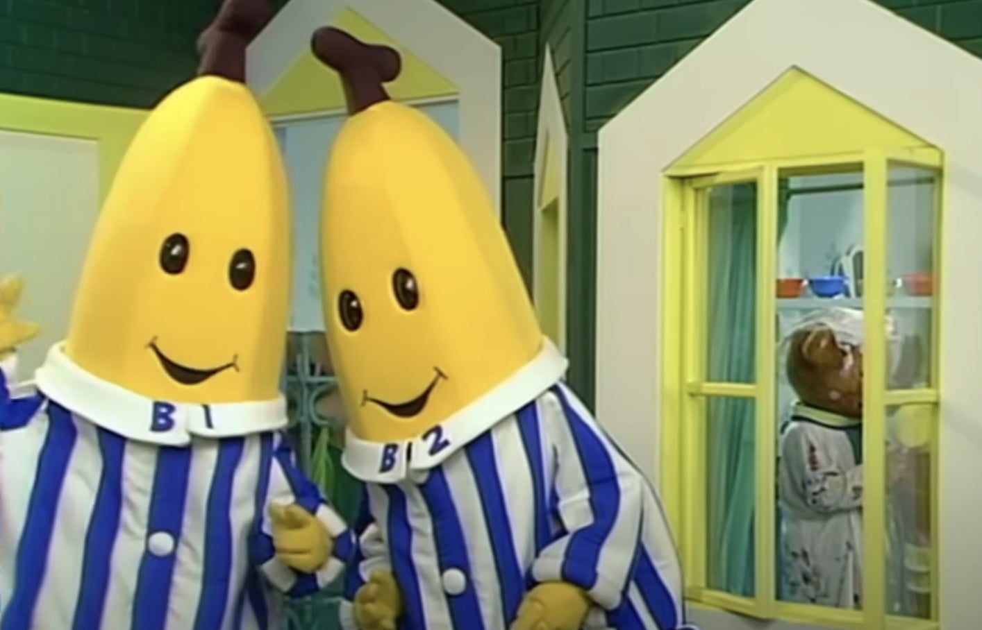 Two people in banana costumes with striped pajamas, labeled B1 and B2, smile indoors. A figure looks through a window in the background