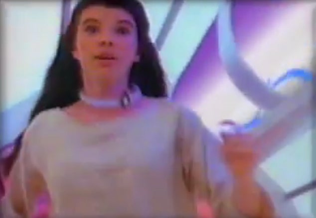 Person in a futuristic setting wearing a light, flowing outfit with long hair. Background features curved, abstract structures
