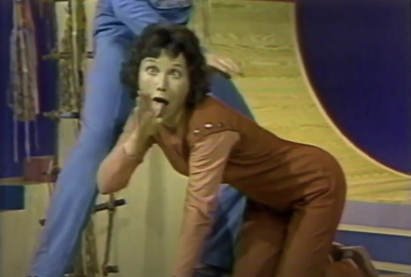 Person in vintage TV show scene, wearing a long-sleeved jumpsuit, posed expressively on set