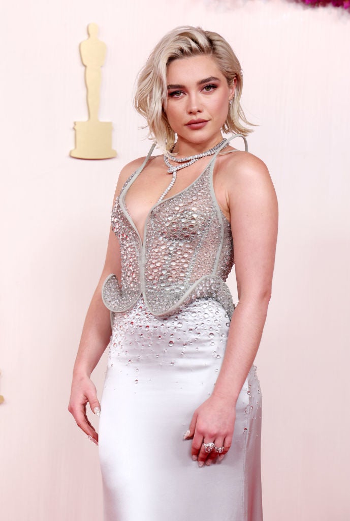 Person in an elegant, embellished gown with a flowing, metallic top and fitted skirt, posing on a red carpet with an award statue in the background
