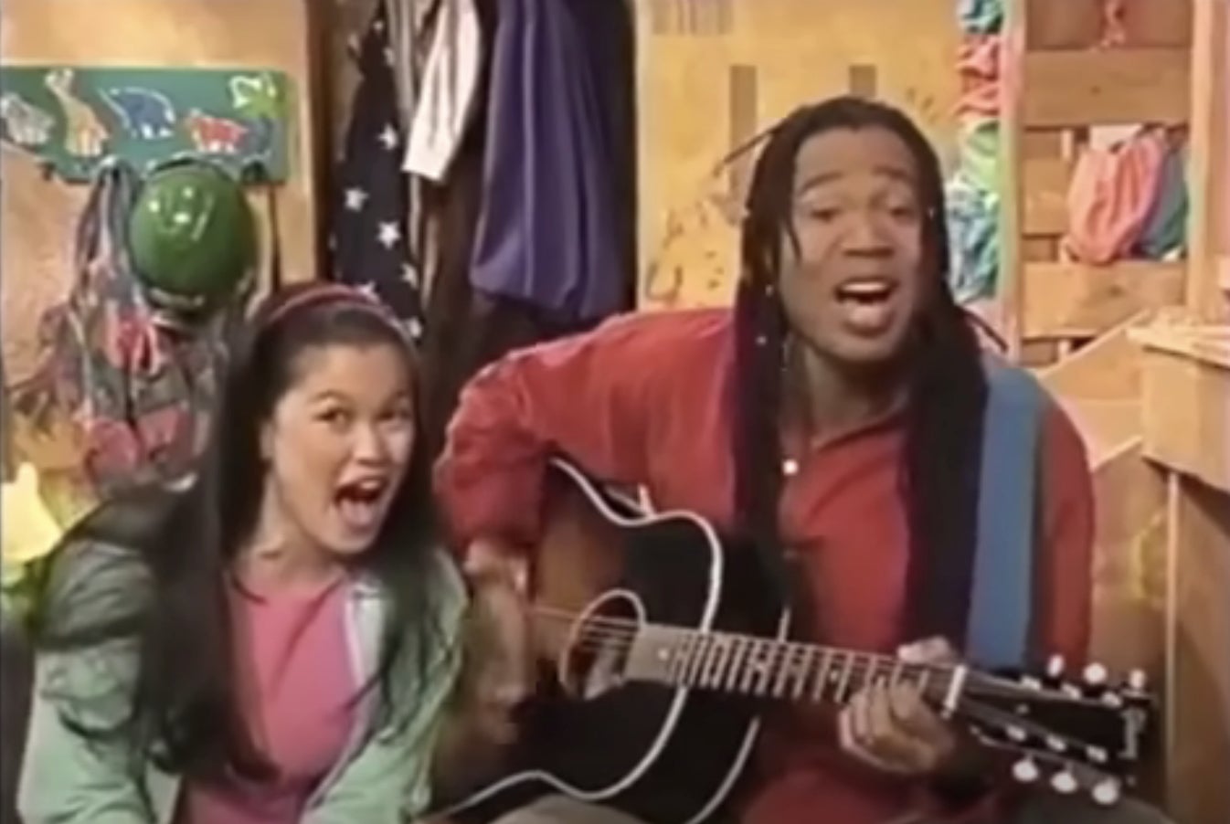 Two people, one playing a guitar, sing enthusiastically in a setting with colorful clothes and helmets in the background