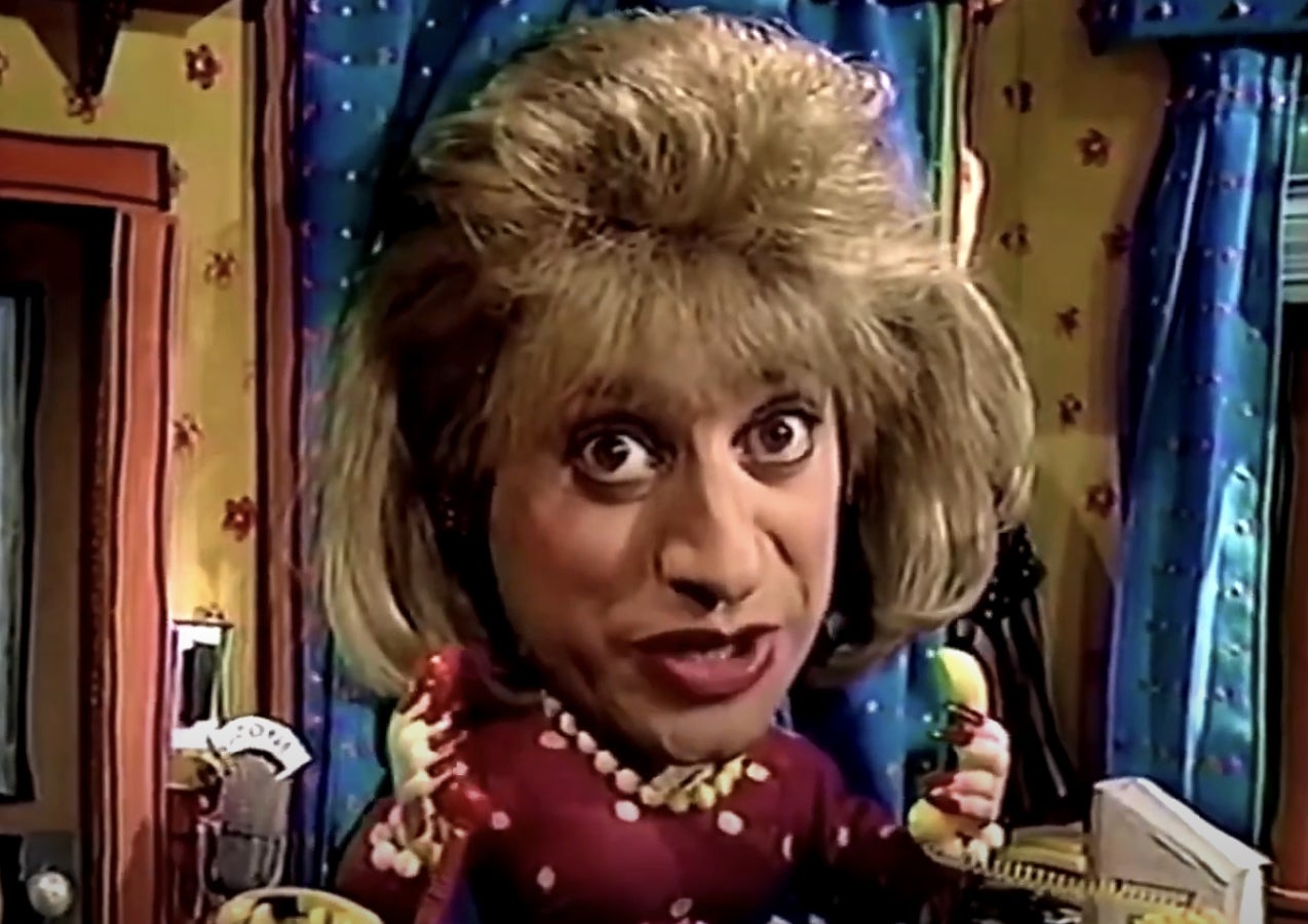 Person with a puppet-like costume, exaggerated makeup, large hair, and a polka dot outfit, possibly from a TV or comedy show scene