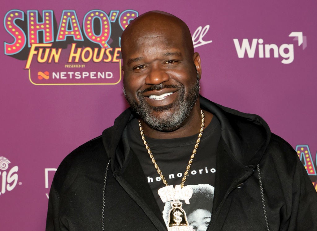 Man smiling at an event backdrop, wearing a black hoodie and graphic T-shirt with a gold chain necklace