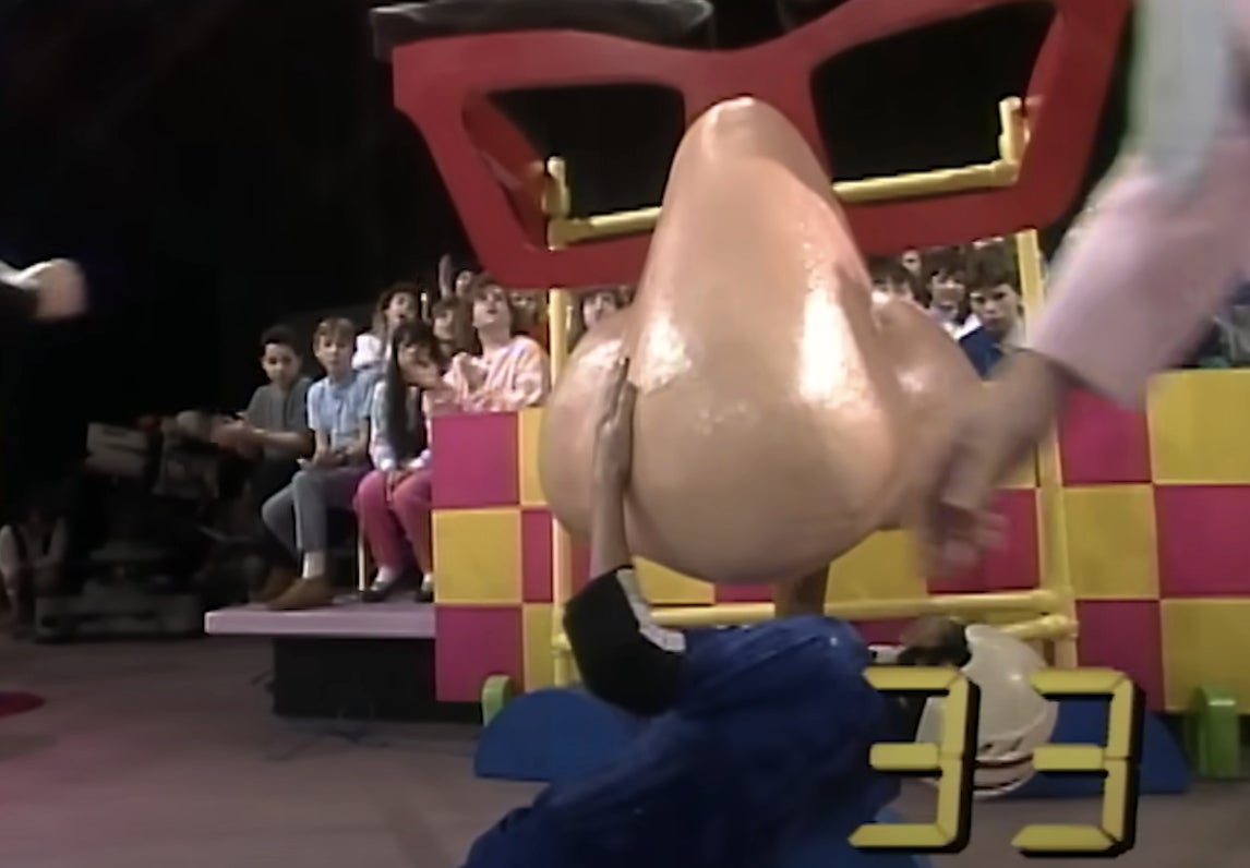 Game show set with giant nose prop; audience watches. Contestant reaches inside as part of a challenge. Timer shows 33 seconds remaining