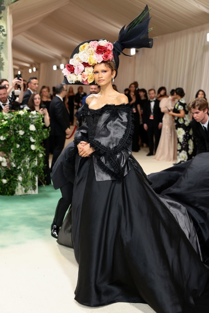 Person in elegant black gown and ornate floral hat on a fashion event red carpet, surrounded by attendees