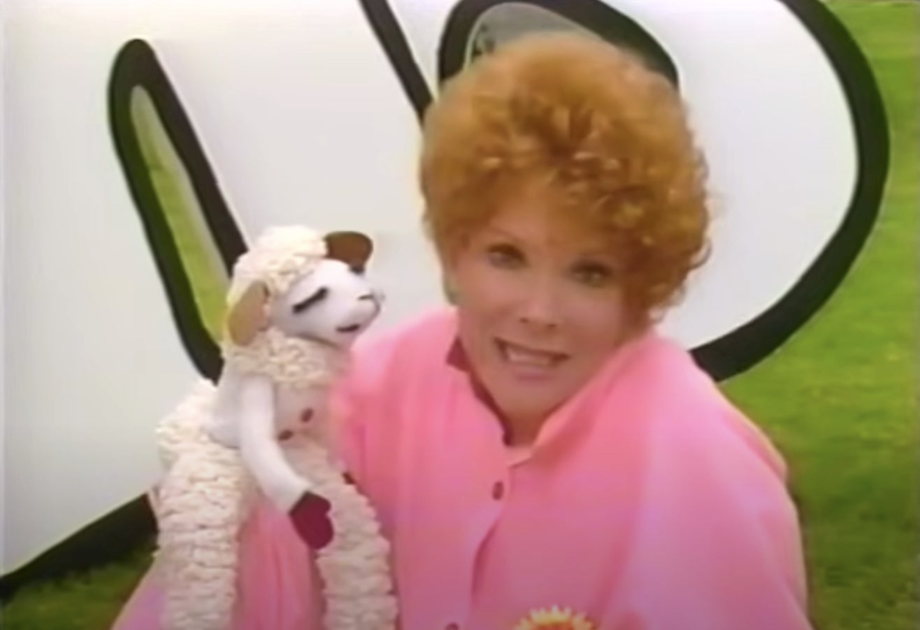 A person with curly hair holds a lamb puppet, standing in front of large letters outdoors