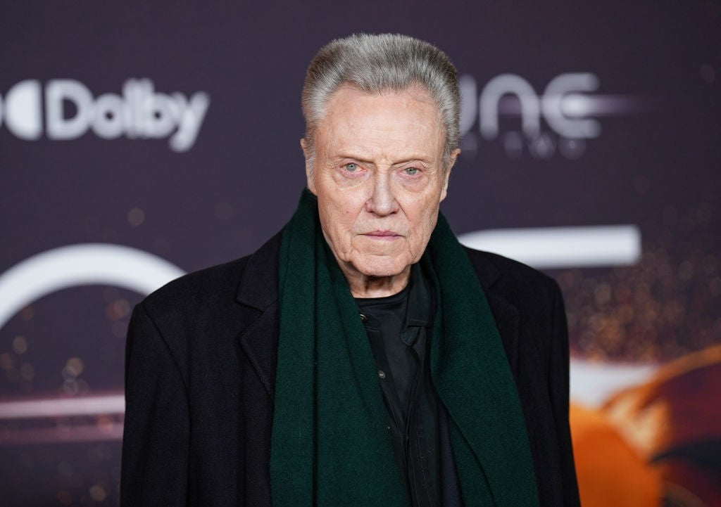 Person on red carpet in a dark coat and scarf, standing in front of a backdrop featuring logos and abstract patterns