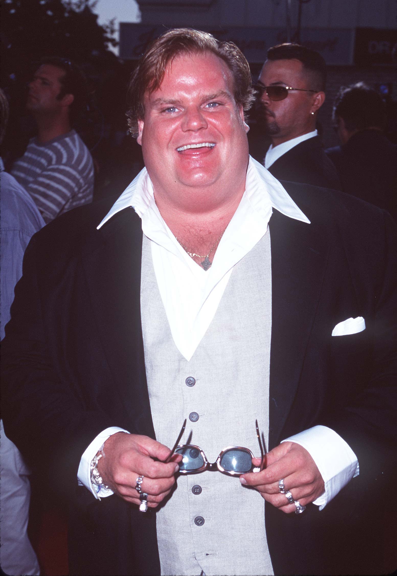 Person smiling, wearing a suit with an open-collared shirt and holding sunglasses in hand at an event