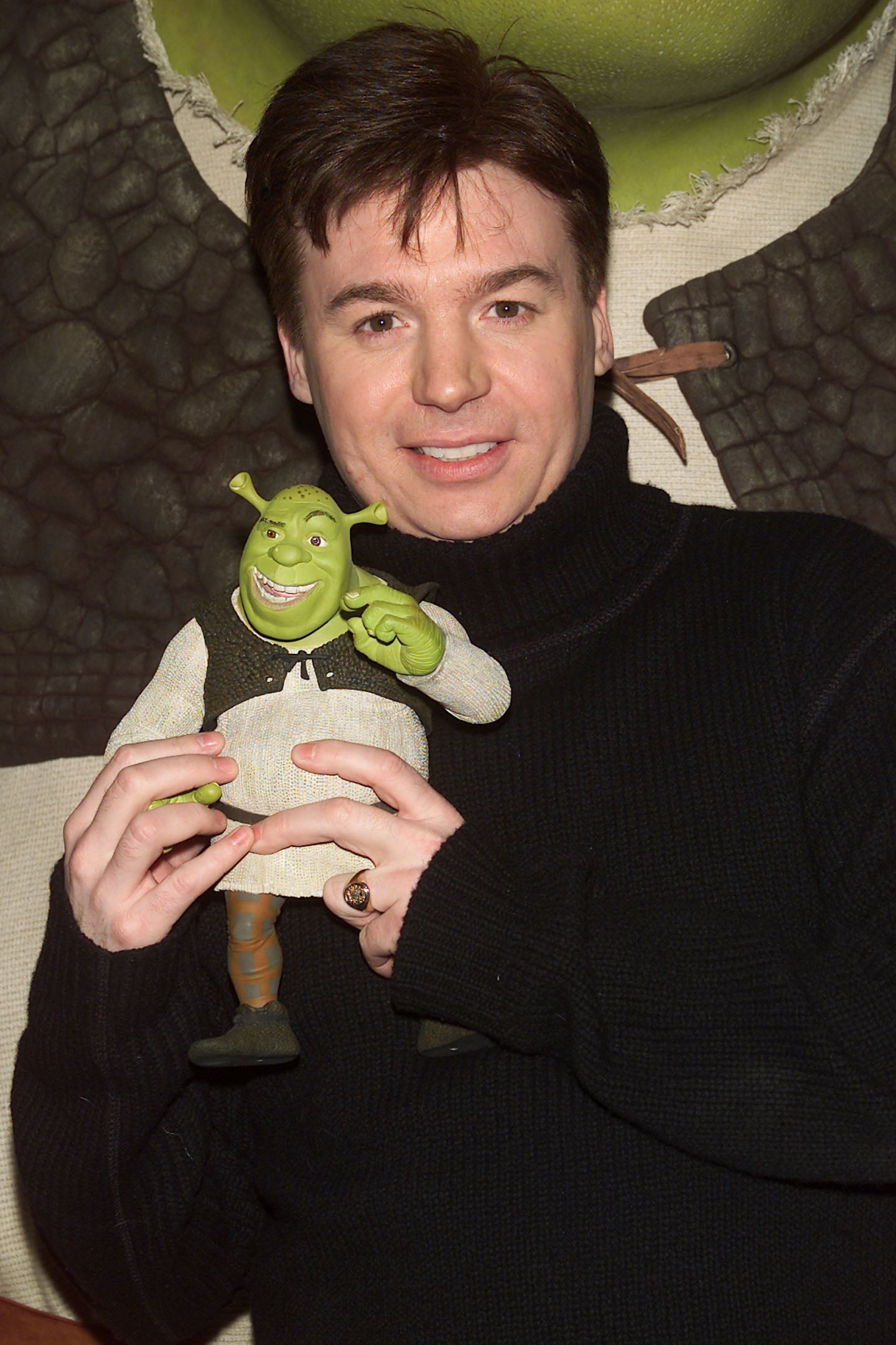 Person holding a Shrek doll, wearing a black turtleneck sweater, standing against a Shrek-themed background