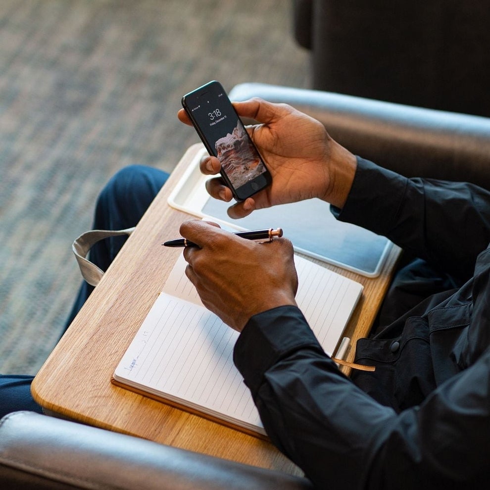 Person seated with a notebook and pen, holding a smartphone displaying a scenic image. Appears focused, possibly planning or shopping online