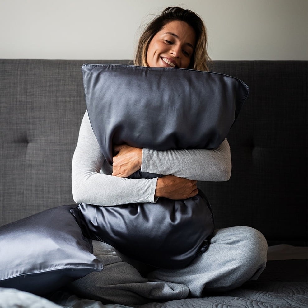 A person sits on a sofa, wearing comfortable loungewear, smiling, and hugging a silky pillow