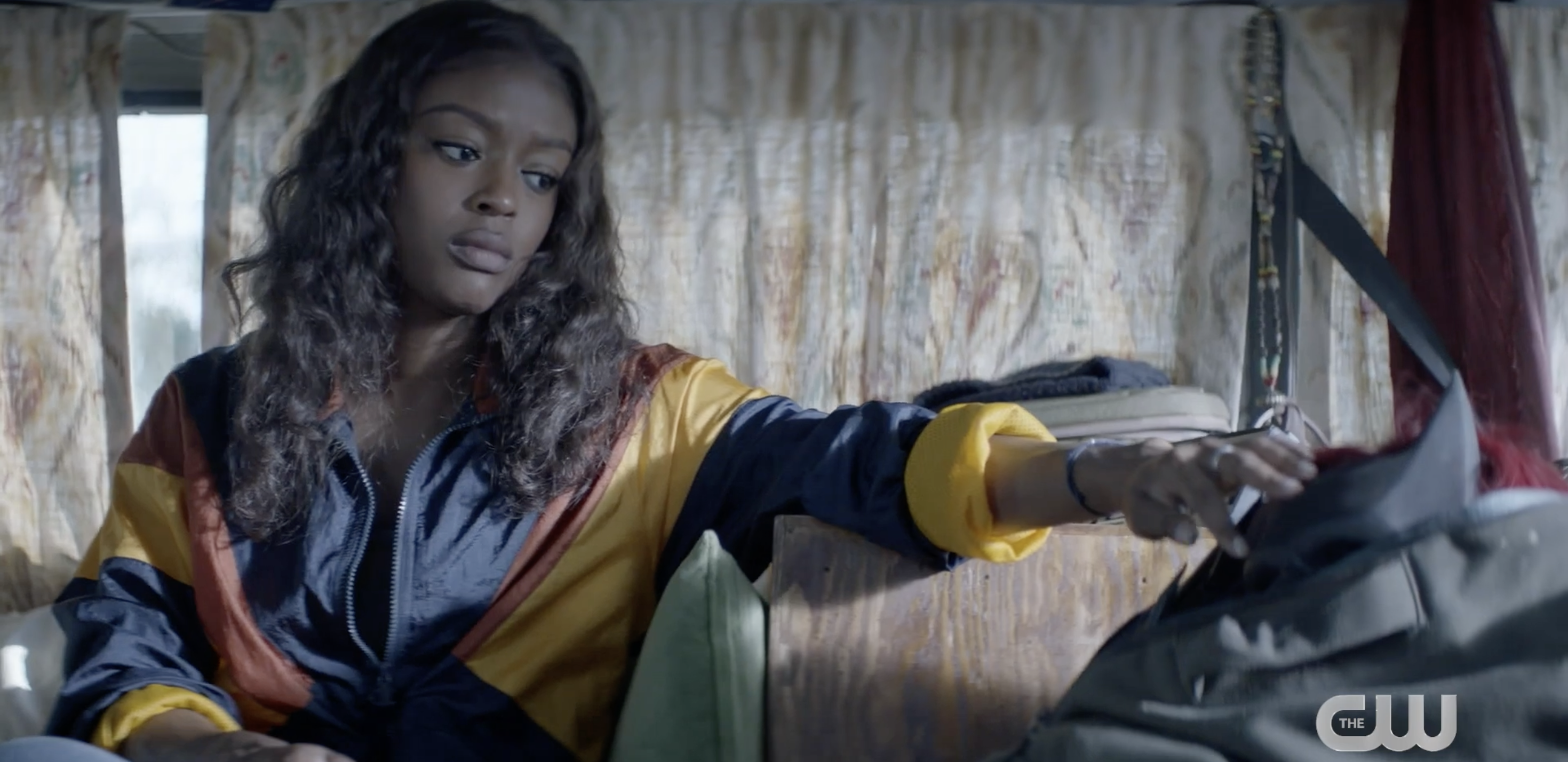 A woman in a retro-style jacket sits inside a vehicle, reaching towards a backpack with a focused expression