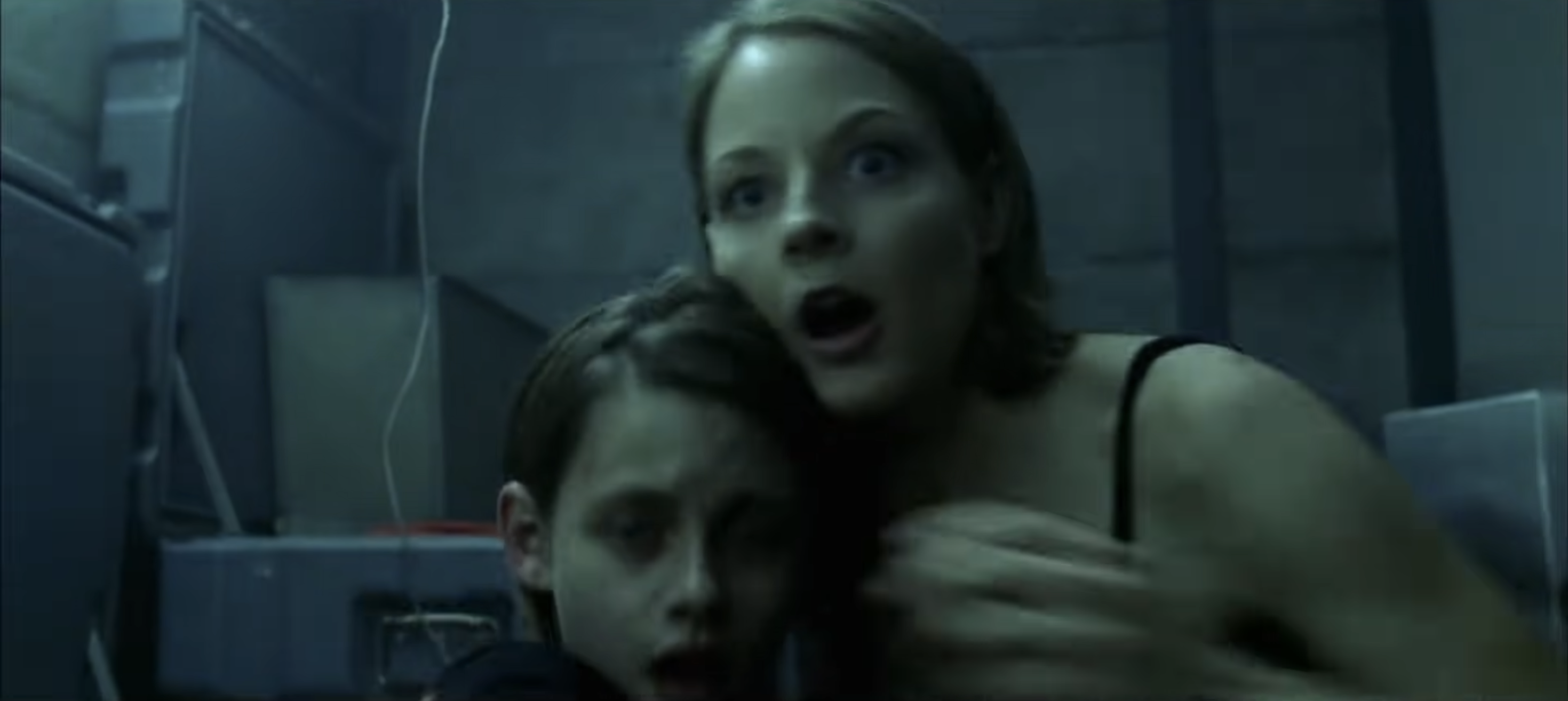 Two people huddle in fear, looking startled, inside a dimly lit room