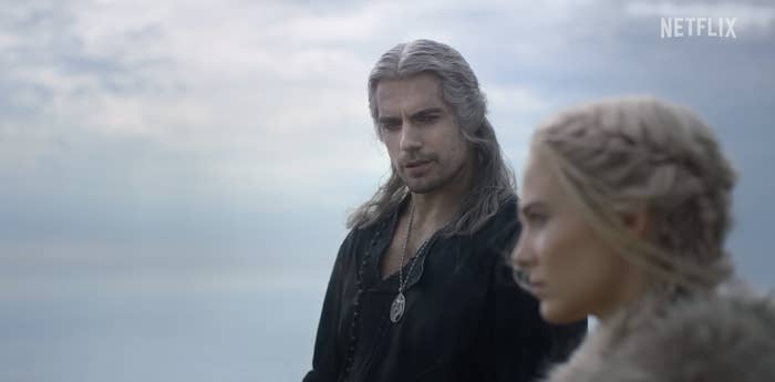 A person with long white hair and a medallion looks at another person with braided hair in a fur cloak, both standing against a cloudy sky