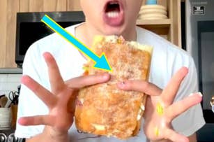 Person holding a large sandwich with vibrant ingredients, showing an open mouth with excitement, inside a modern kitchen