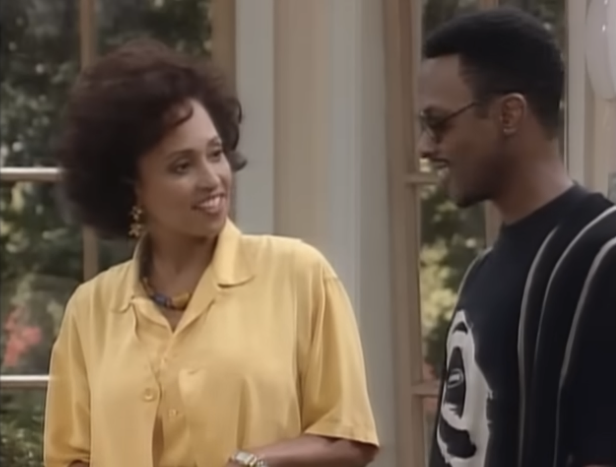 Two people smiling at each other indoors; one wears a yellow blouse, the other a black shirt with a bold graphic design