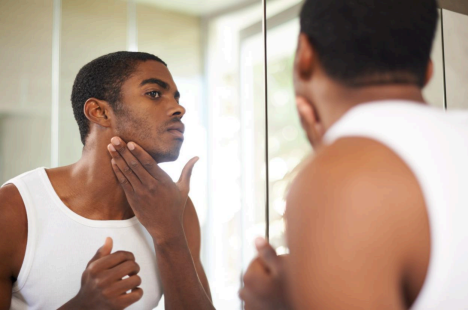 Man in a white tank top examining his face in a bathroom mirror, touching his cheek thoughtfully