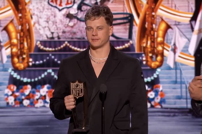 Joe Burrow holding an NFL award, dressed in a formal black suit and pearl necklace, stands on stage with an ornate background featuring floral elements
