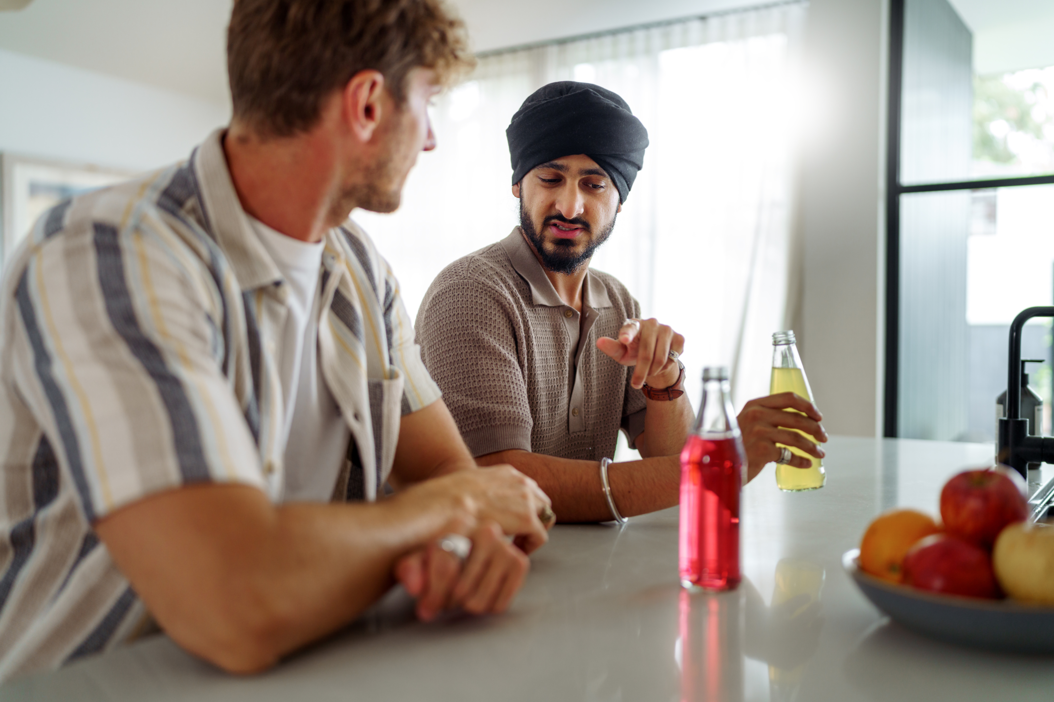 To menn sitter ved en kjøkkenbenk. Den ene har på seg en stripet skjorte og den andre har en turban. De diskuterer mens de holder drikke, med frukt i nærheten