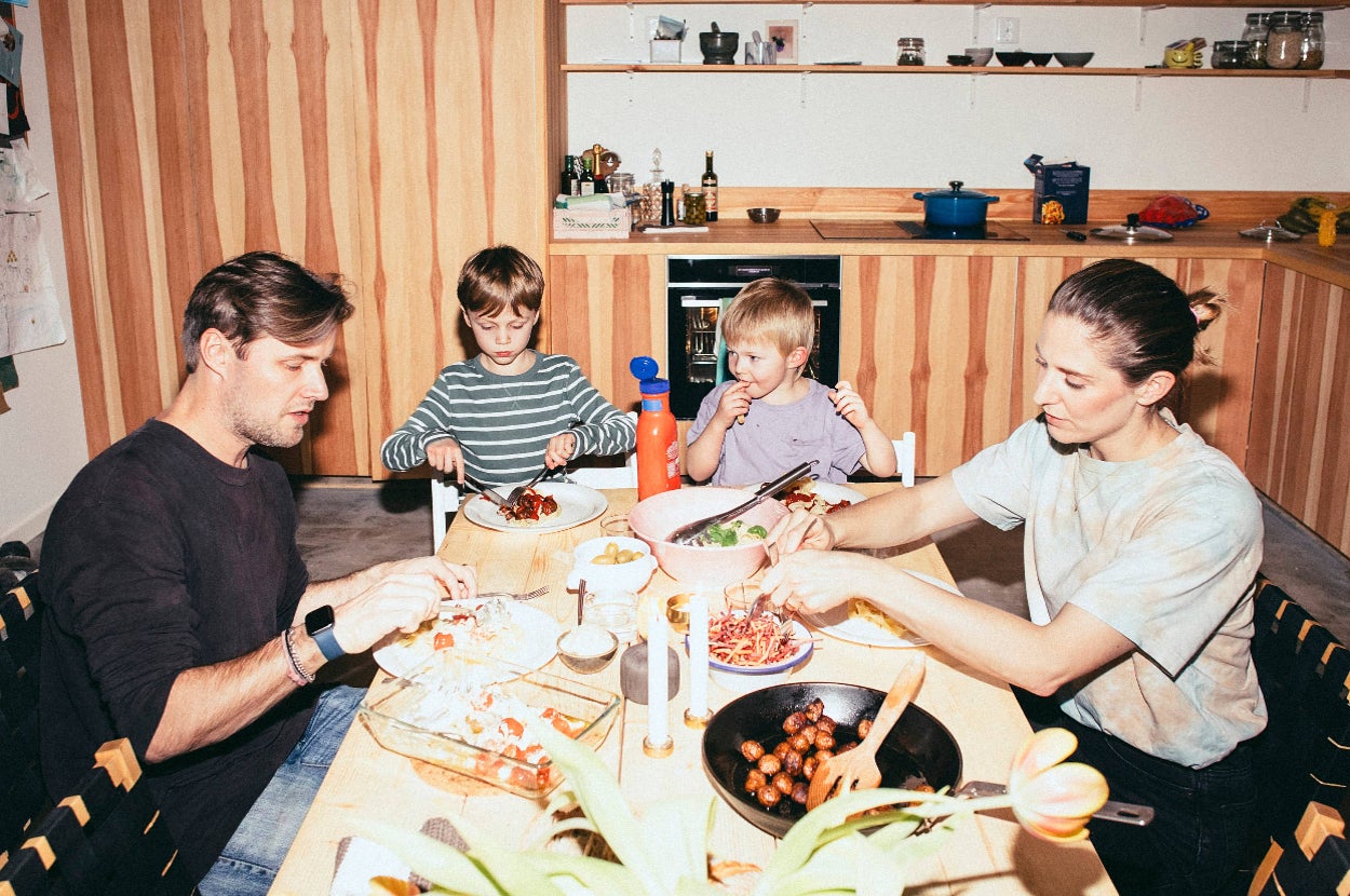 Family of four sharing a meal at a wooden dining table in a modern kitchen. Parents and two young children are enjoying various dishes together