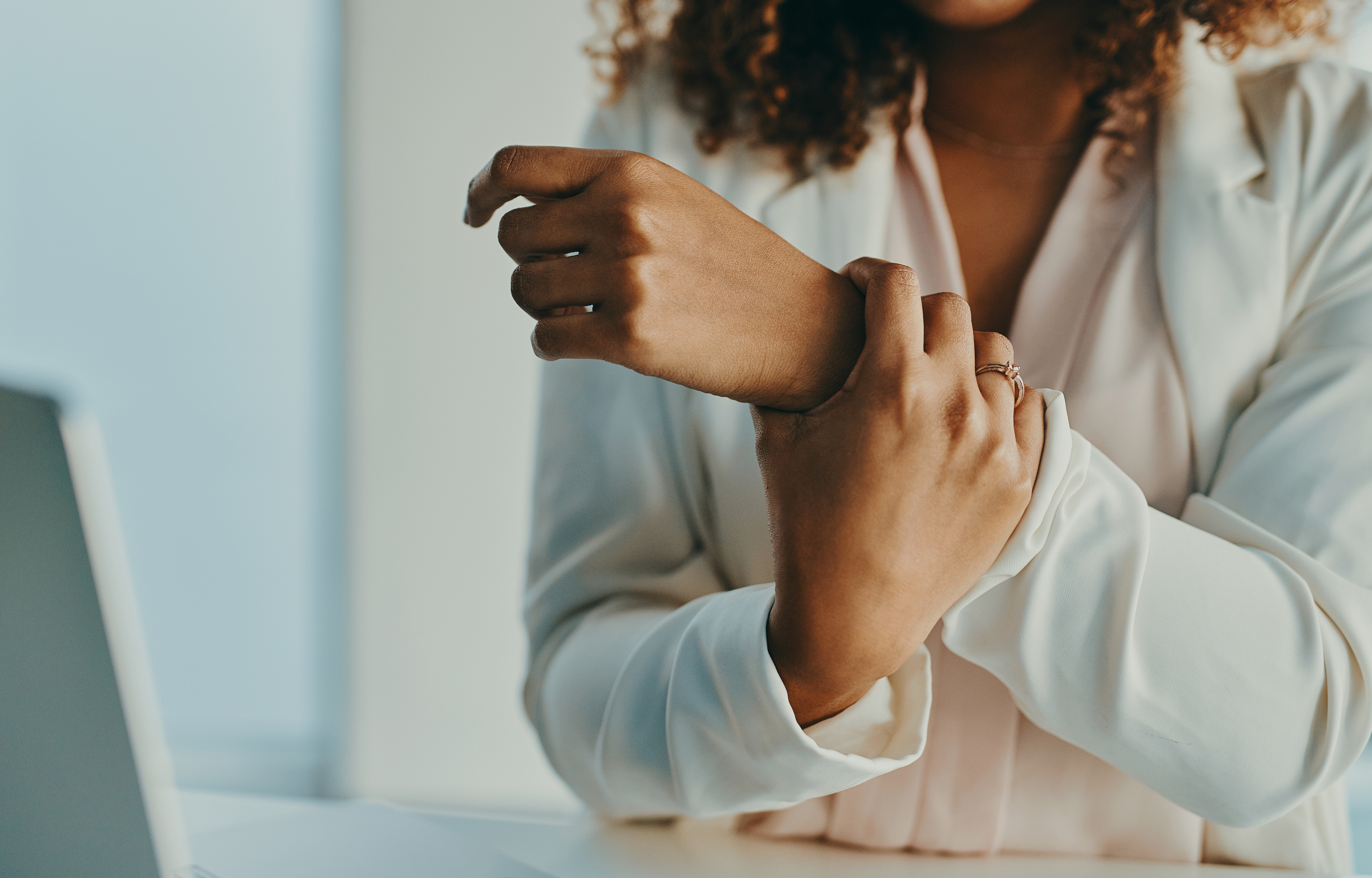 Person i en hvit skjorte som forsiktig holder håndleddet, sittende ved et skrivebord, og antyder et øyeblikk av refleksjon eller mindfulness