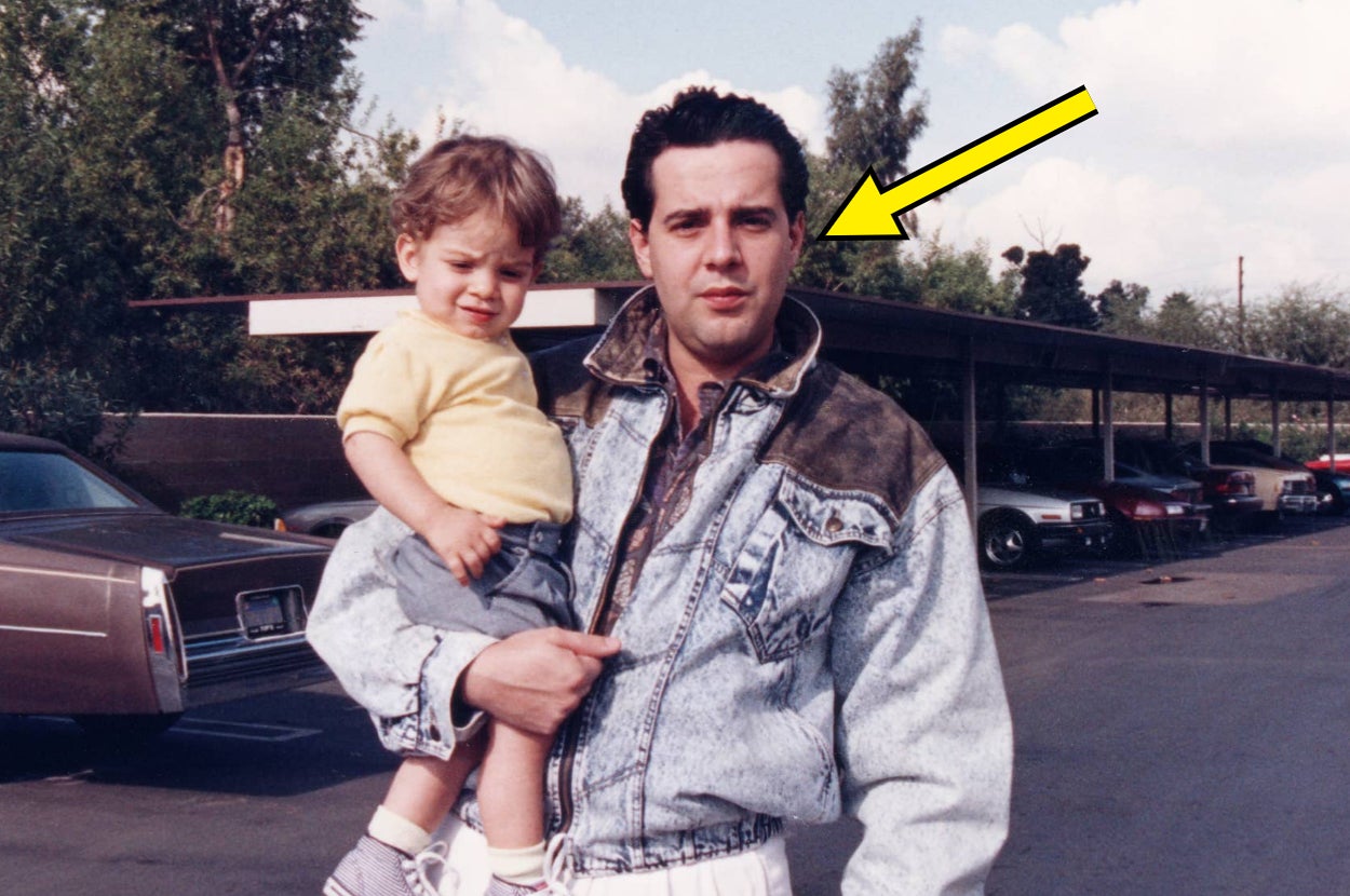 A man in a denim jacket holds a child in a parking lot, looking at the camera. A yellow arrow points to the man