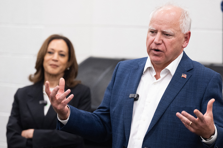 室内でスーツ姿の男性がスピーチをしている。背景に別のスーツ姿の人物が立っている。