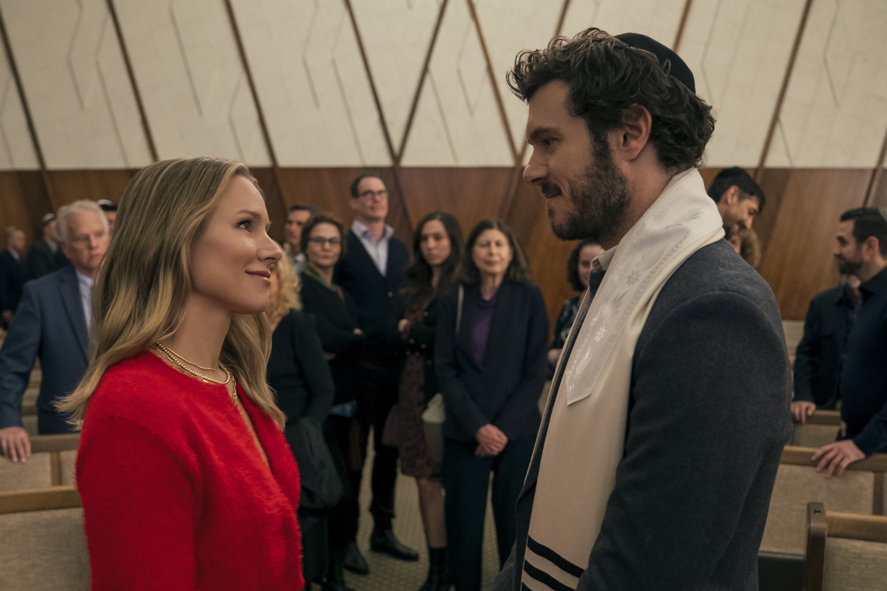 Two people smile at each other in a formal setting, surrounded by an audience. The woman wears a red outfit; the man has a prayer shawl