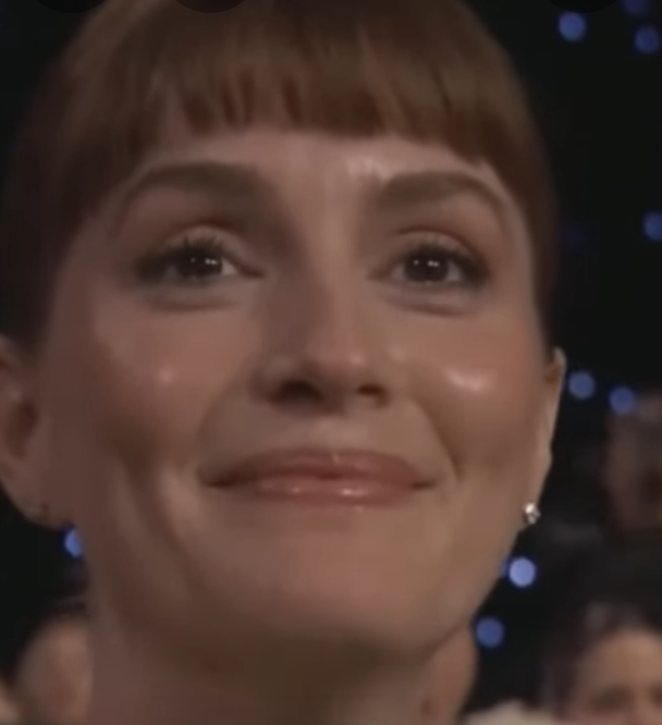 Person at a formal event, smiling gently, with short hair and wearing earrings