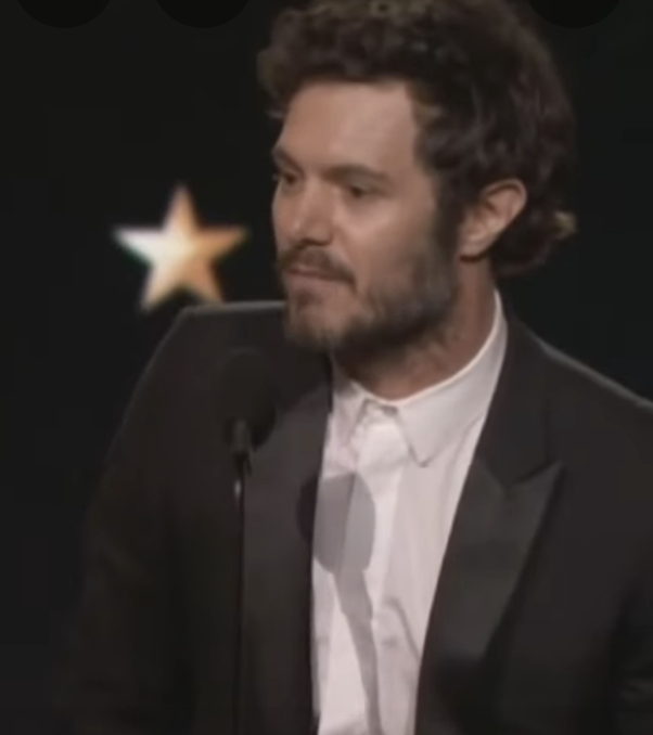 Person in a suit speaks at an event. A star is visible in the background