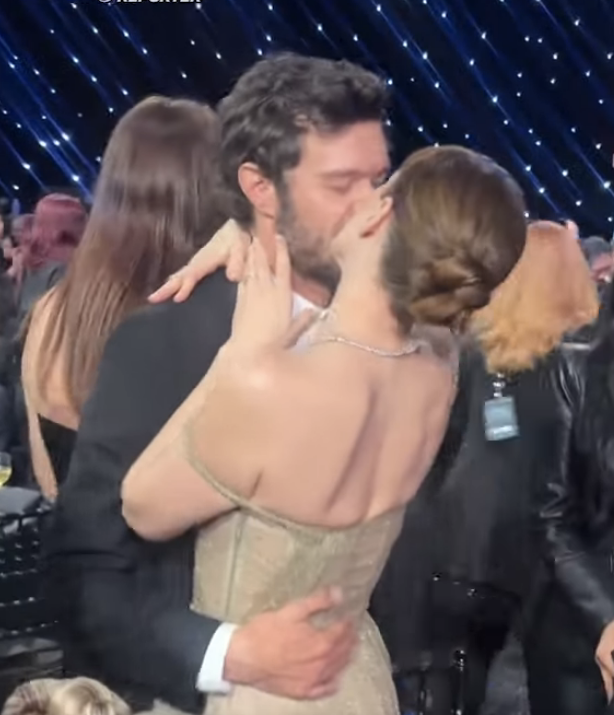 Two people embrace and kiss at a formal event. One wears a strapless gown, and the other is in a suit