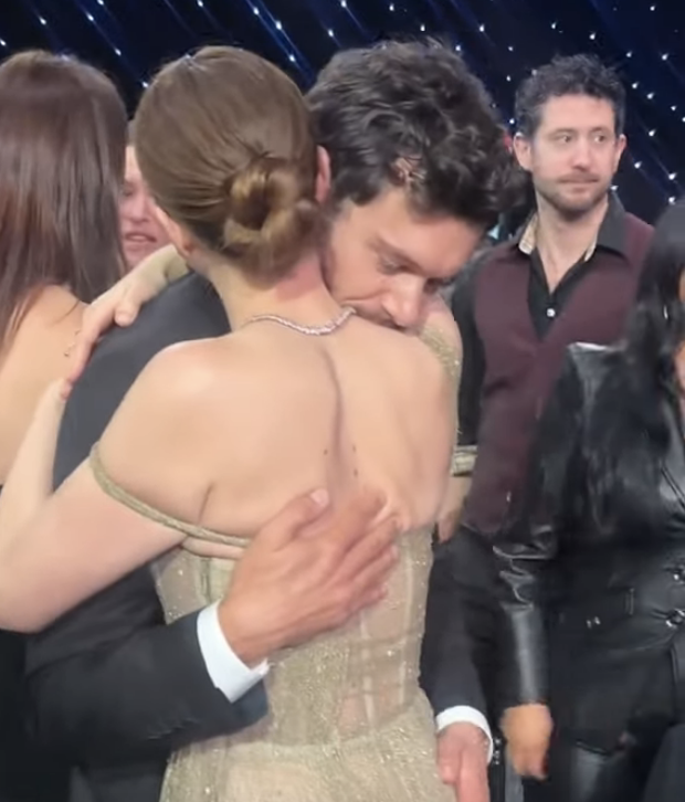 Two people embrace on a red carpet, one wearing a formal gown. Others are in the background
