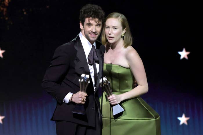 Two people on stage holding awards; one in a tuxedo, the other in a stylish strapless dress