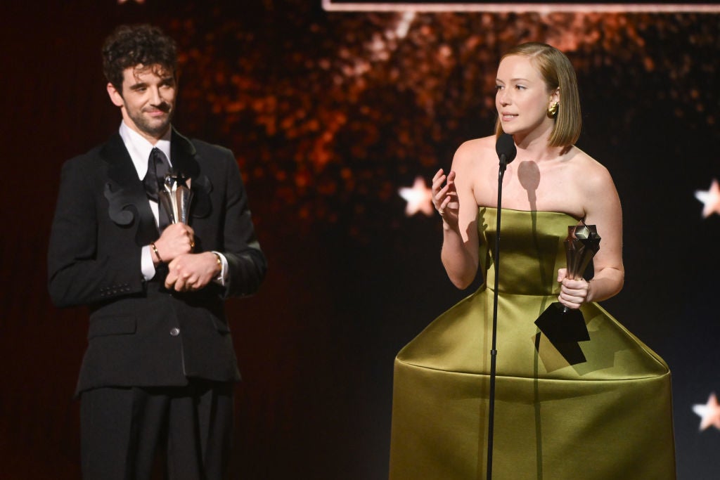 Two people on stage at an awards show. The woman wears a structured, geometric dress; the man is in a classic suit, both holding trophies