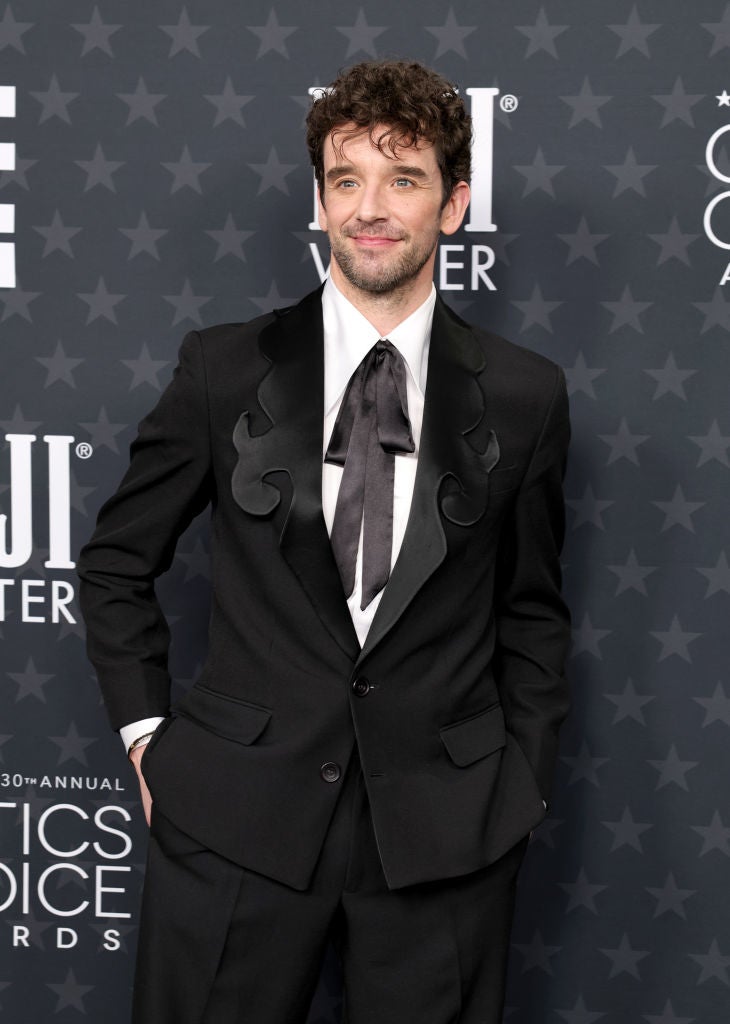 Person in a stylish black suit with unique lapel design and large bowtie, posing on a star-patterned background at an awards event