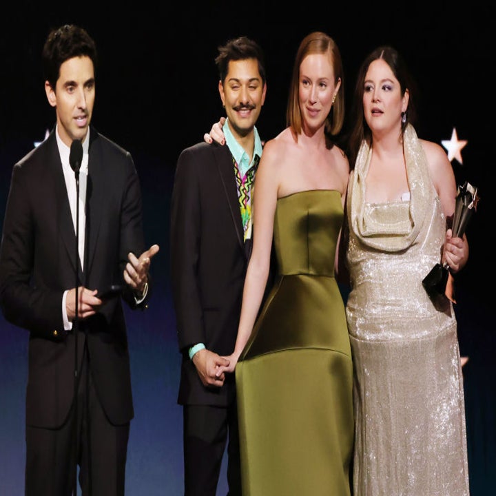 Four people on stage, one in a suit speaking at a podium, others in formal attire holding an award trophy with expressions of joy and camaraderie