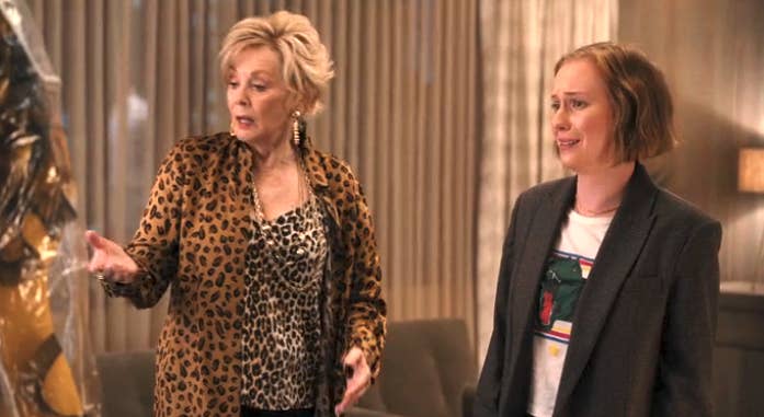 Two women in a room, one in a leopard print outfit, the other in a blazer and graphic tee, are engaged in a conversation, showing animated expressions