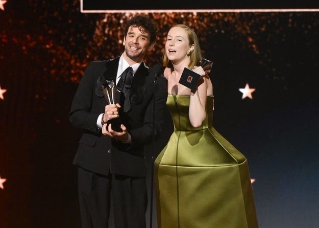 Two people on stage at an award event. The person on the left wears a formal suit; the person on the right wears a structured, stylish dress
