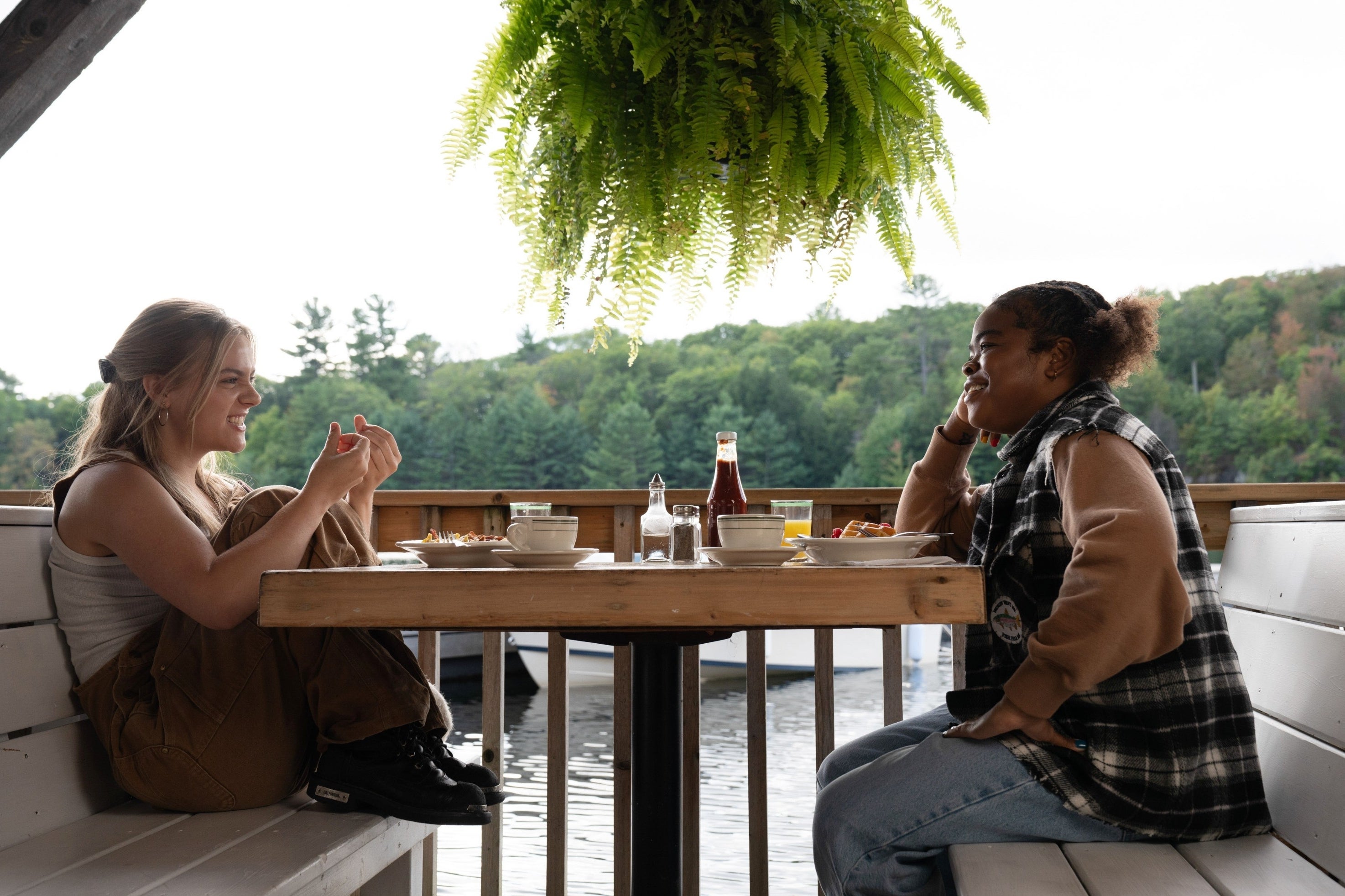 Two people enjoying a meal together at an outdoor table by a lake, with greenery in the background. One person is wearing brown overalls, the other a plaid jacket