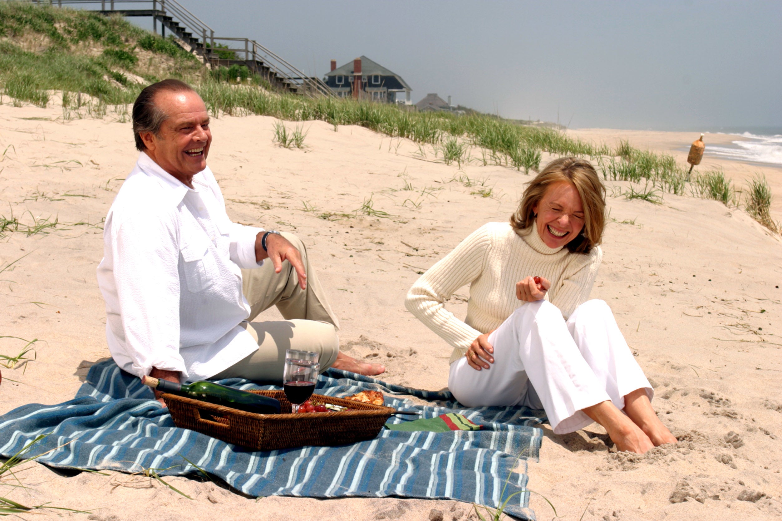 Two people enjoying a picnic on a beach blanket, laughing and relaxing by the shore. One has a basket with food and drinks